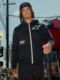 Alpinestars Transverse Hoodie, schwarzes Kapuzenfleece mit Reißverschluss vorne, weißer Reißverschluss und weißes Astars-Logo auf der Brust, „One Goal One Vision“ Textaufdruck, lässiger Streetwear-Stil, getragen von einem Mann in einer urbanen Außenumgebung