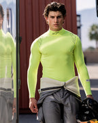 Alpinestars ZX Evo V3 Langarm-Top, Auto-Rennsport-Basisschicht, Gelb Fluo, getragen von einem männlichen Fahrer mit um die Taille gebundenem grauen Rennanzug, hält einen schwarzen Helm, Alpinestars-Logo an Hals und Schultern, technische feuchtigkeitsableitende Motorsport-Ausrüstung