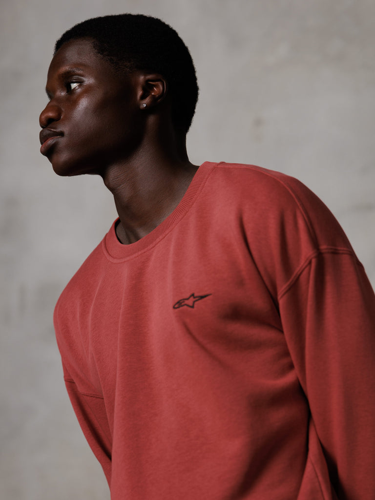 Alpinestars Essentials Ageless Crew, ziegelrotes Rundhals-Sweatshirt, von einem männlichen Model im Seitenprofil getragen, kleines schwarzes Stern-Logo auf der Brust, entspannte Passform, hochwertige Fleece-Streetwear