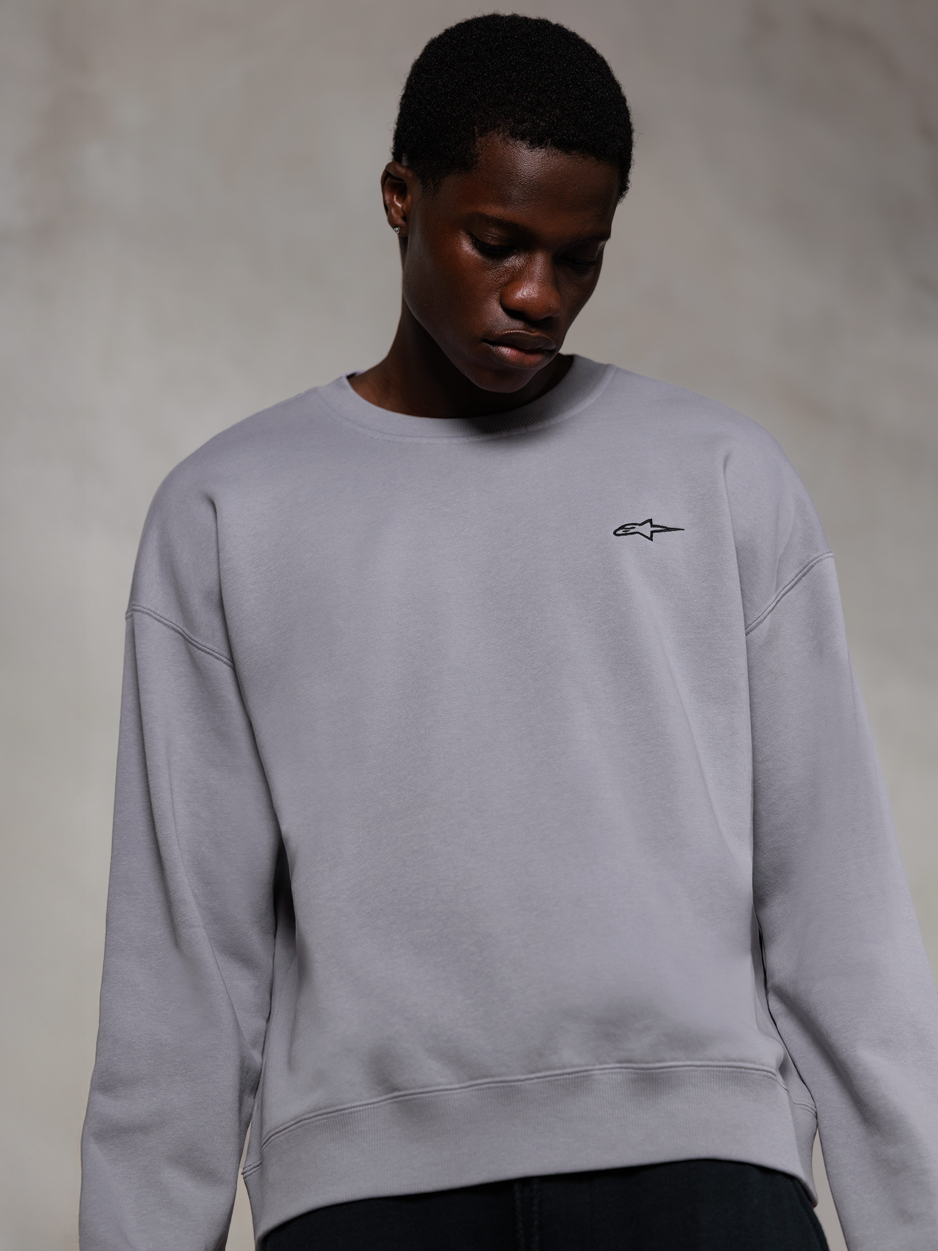 Alpinestars Essentials Ageless Crew, hochwertiges Rundhals-Sweatshirt in Silber, mit einem kleinen schwarzen Alpinestars-Logo auf der Brust, von einem Model getragen, lässige Lifestyle-Streetwear mit weicher Fleece-Textur.