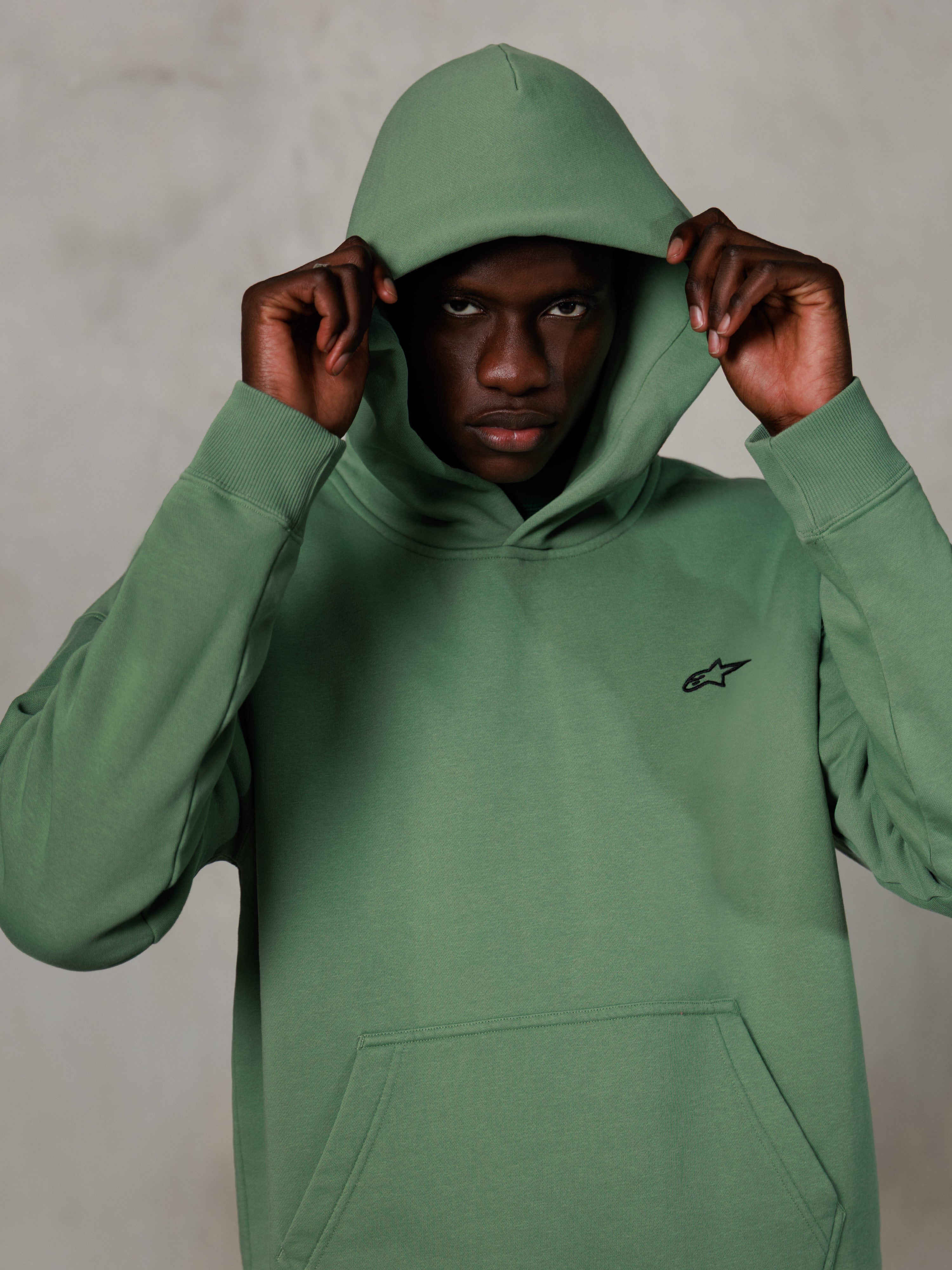 Alpinestars Essentials Ageless Hoodie, lässige Streetwear, Kaktusgrün mit kleinem schwarzem Logo auf der Brust, Modell trägt den Hoodie mit aufgesetzter Kapuze, Kängurutasche vorne, entspanntes, vom Motorsport inspiriertes Design
