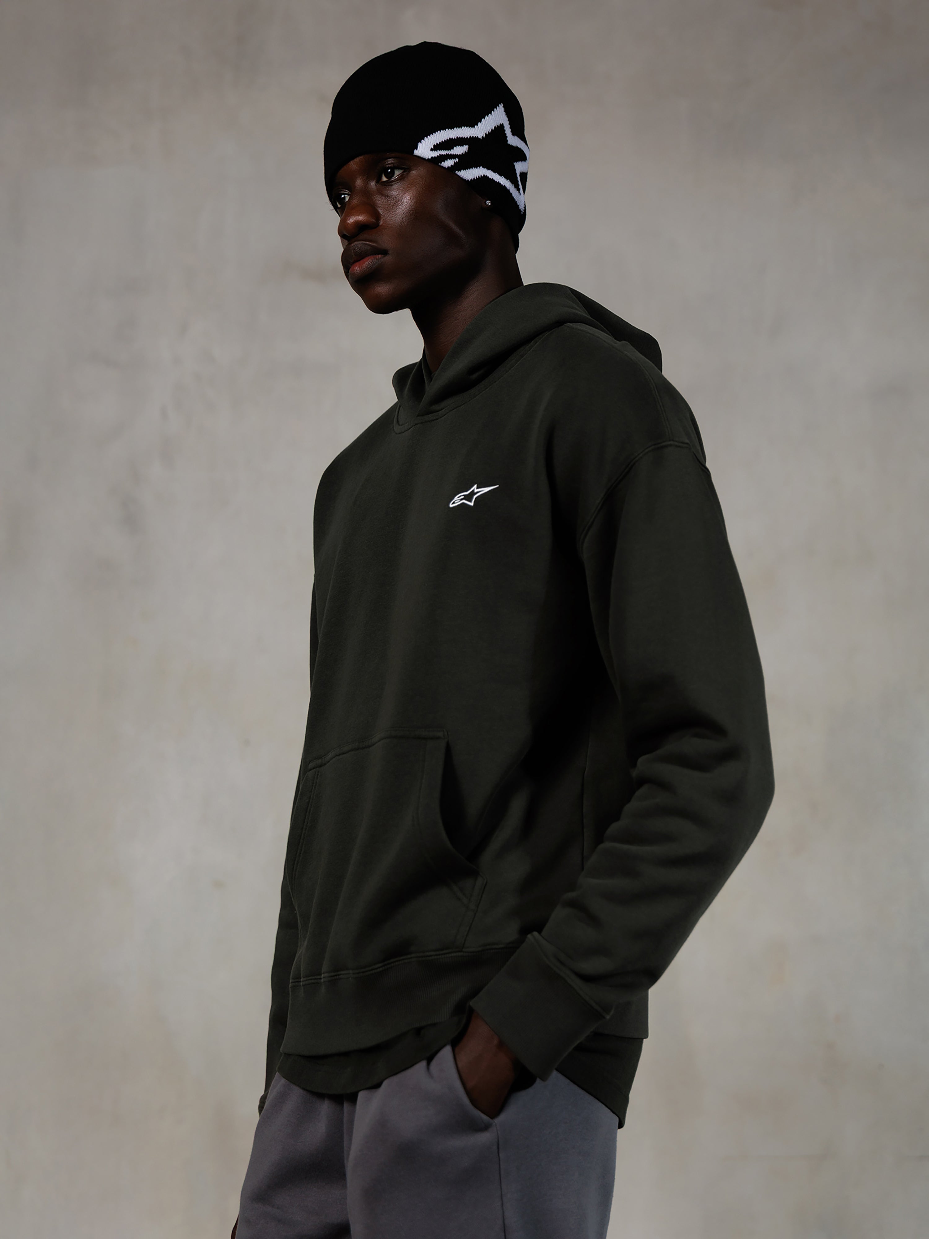 Alpinestars Essentials Ageless Hoodie, Off Black, männliches Model in lässiger Streetwear, dunkelgraues Fleece mit Stone-Wash-Finish und kleinem weißen Stern-Logo, kombiniert mit einer schwarzen Alpinestars Beanie und einer grauen Hose