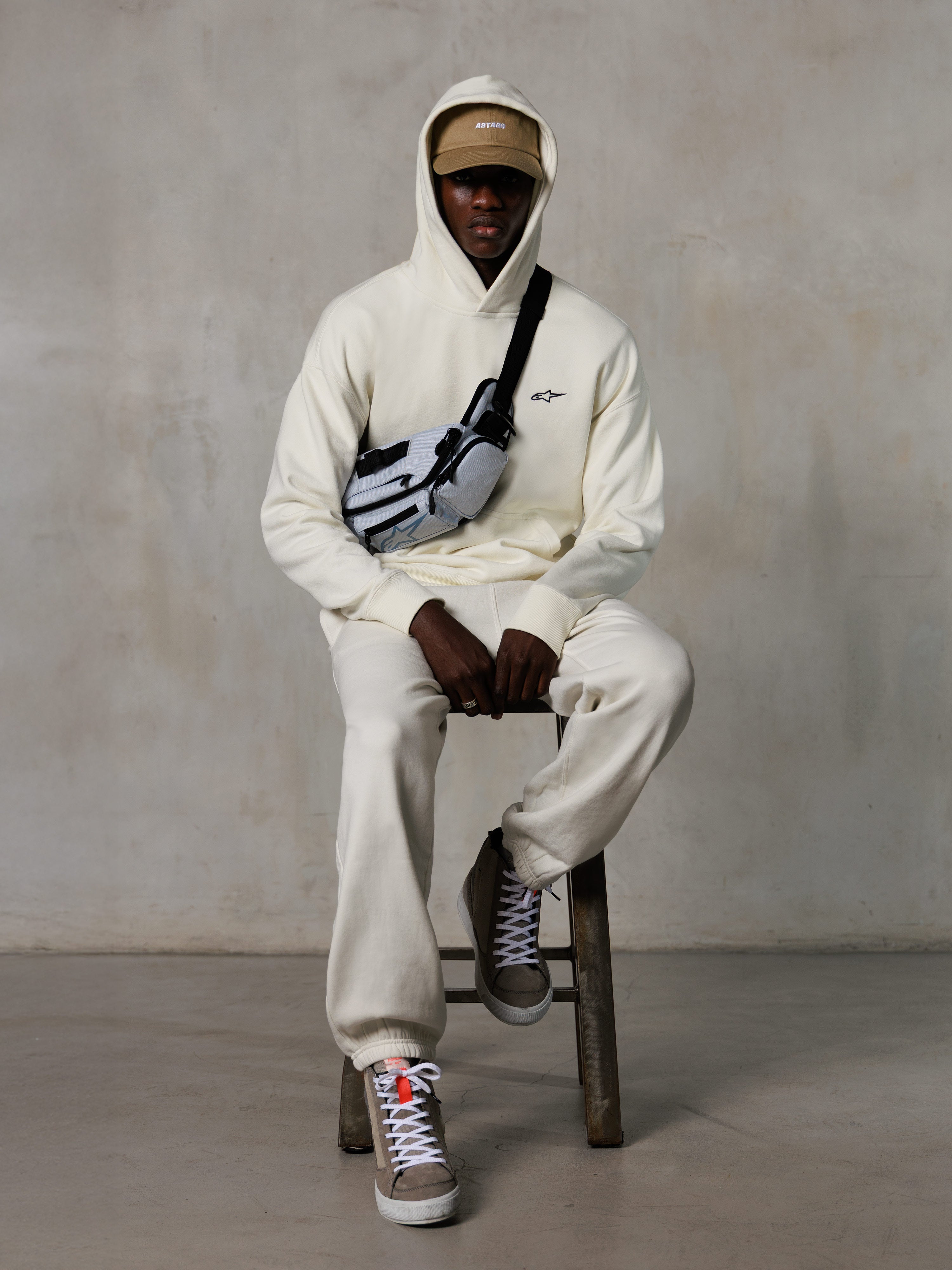 Alpinestars Essentials Ageless Hoodie, Off-White, getragen von einem Modell auf einem Hocker mit passender Fleece-Jogginghose, hellbrauner ASTARS-Cap und Crossbody-Tasche, lässige vom Motorsport inspirierte Streetwear