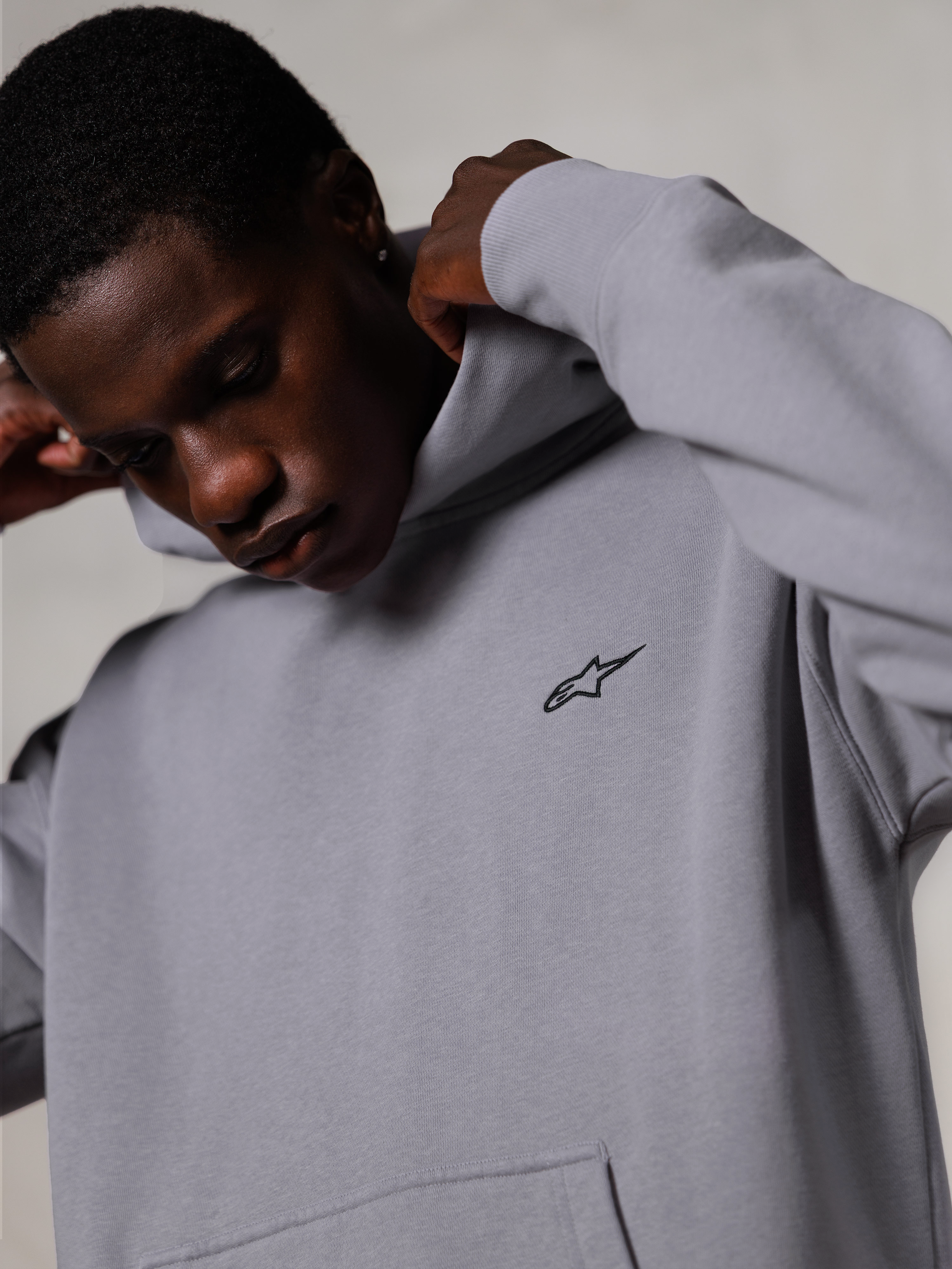 Alpinestars Essentials Ageless Hoodie, Silber, Nahaufnahme eines Mannes, der die Kapuze des grauen Sweatshirts richtet, kleines schwarzes Alpinestars-Logo auf der Brust, hochwertige, vom Motorsport inspirierte Freizeitkleidung