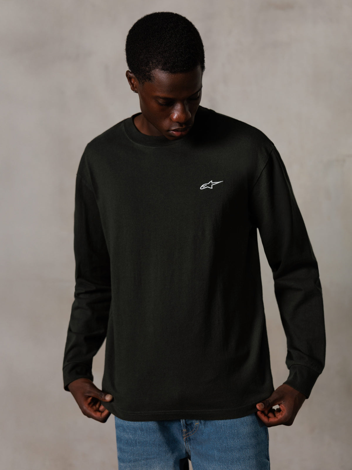 Alpinestars Essentials Ageless LS Tee, Langarmshirt, Off Black, mit einem kleinen weißen Logo auf der Brust, getragen von einem Modell mit blauen Jeans, lässige Lifestyle-Streetwear