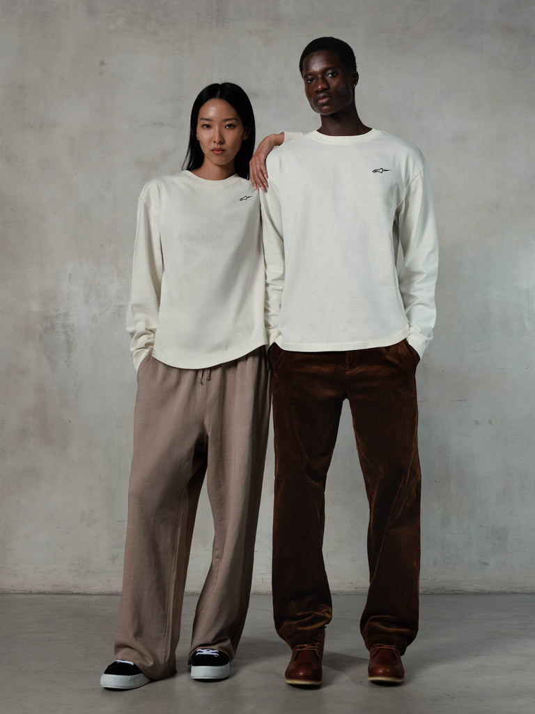 Alpinestars Essentials Ageless LS Tee, Langarmshirt, Off White, getragen von einem männlichen und einem weiblichen Model, kleines schwarzes Alpinestars-Logo auf der Brust, entspannte Passform, kombiniert mit hellbrauner Jogginghose und brauner Cordhose, lässige motorsport-inspirierte Streetwear