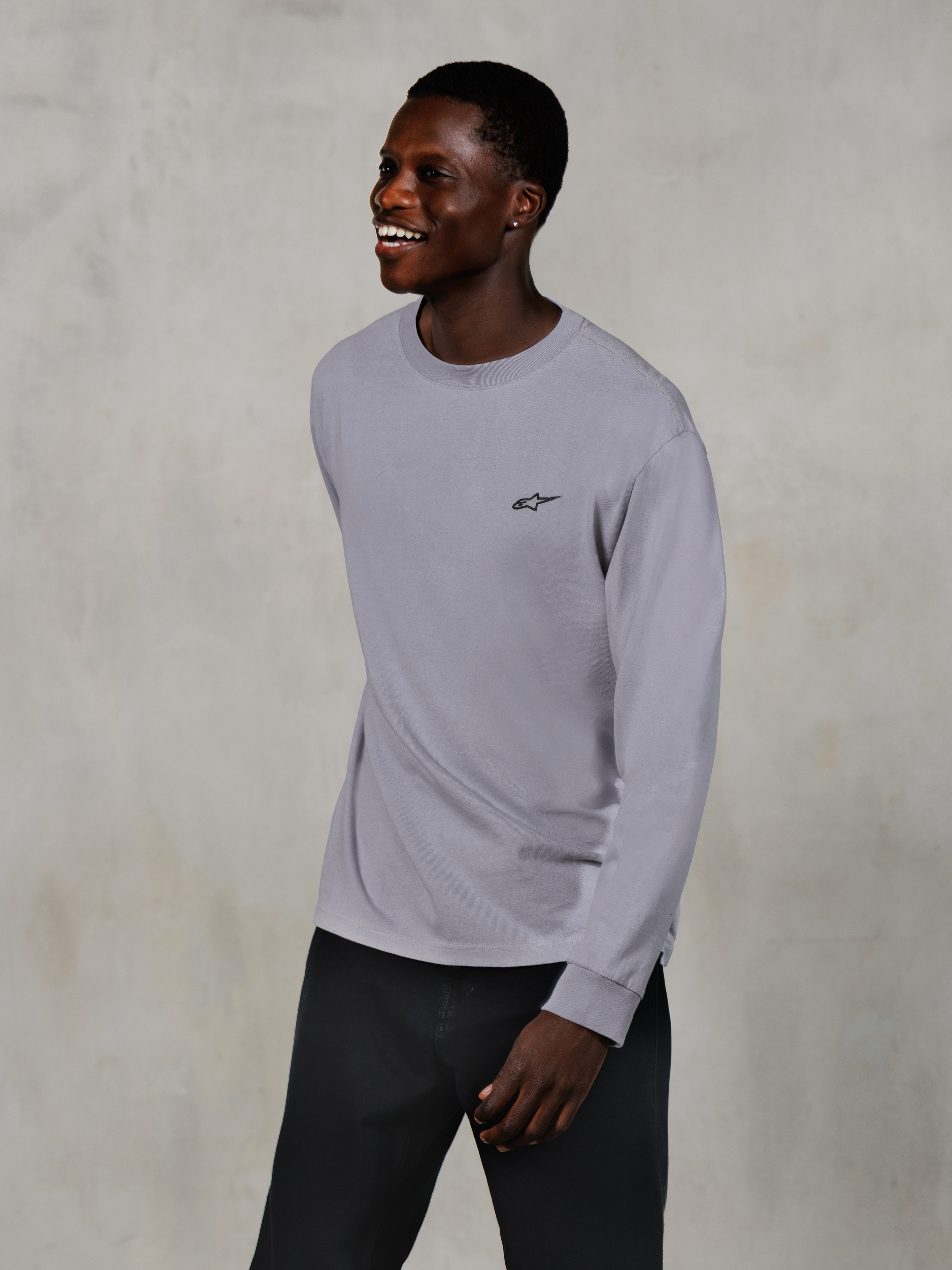 Alpinestars Essentials Ageless LS Tee, Langarm-T-Shirt, Silber, kleines schwarzes Logo auf der Brust, getragen von einem lächelnden Model mit dunkler Hose, entspannte zeitgemäße Passform, lässige Motorsport-Lifestyle-Bekleidung