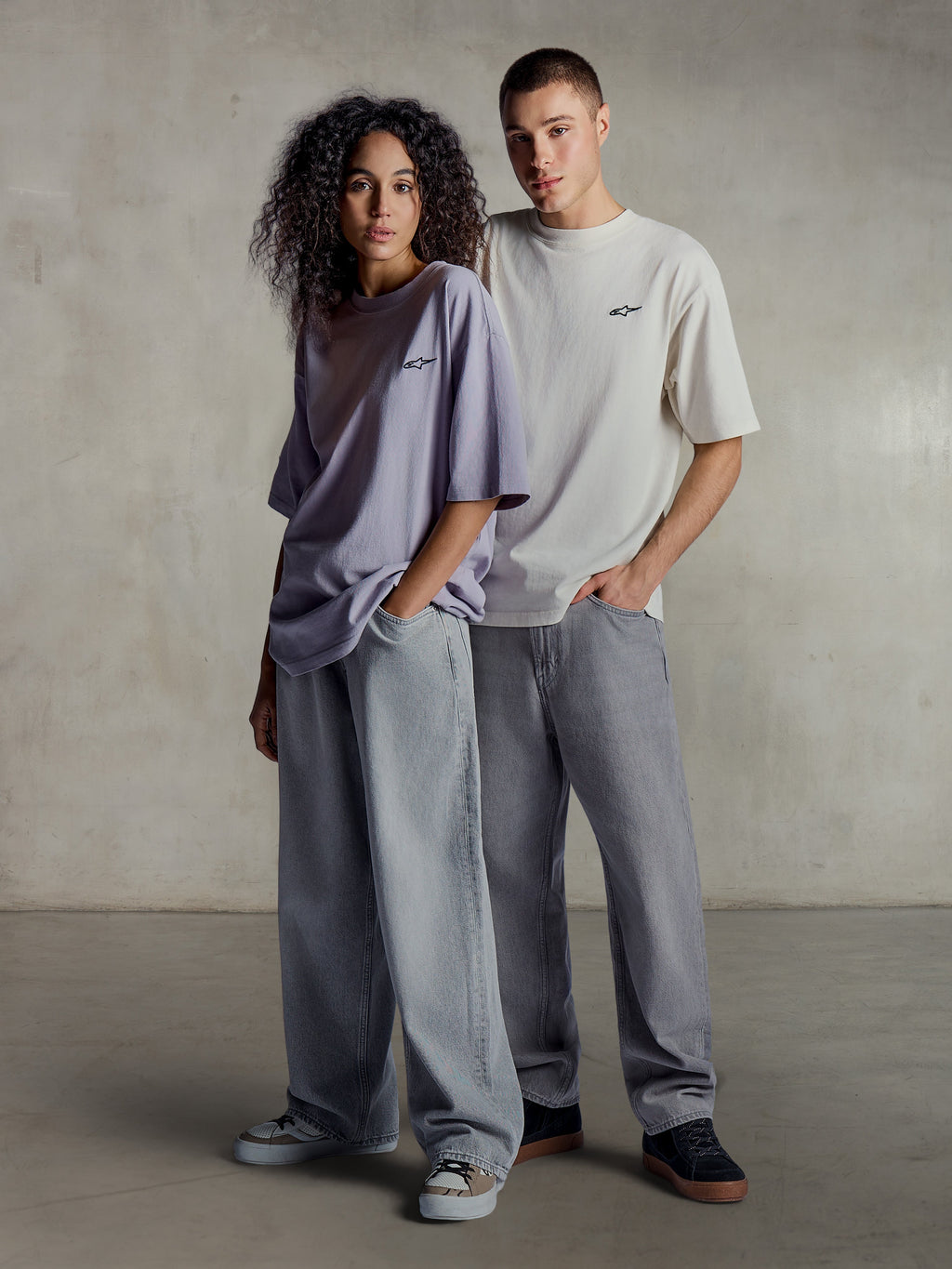 Alpinestars Essentials Ageless SS Tee, Lifestyle-Ansicht von zwei Models in kurzärmeligen T-Shirts in Helllila und Silber, entspannte Passform aus Bio-Baumwolle mit dezentem Brustlogo, kombiniert mit weiten Jeans für einen lässigen Streetwear-Stil.
