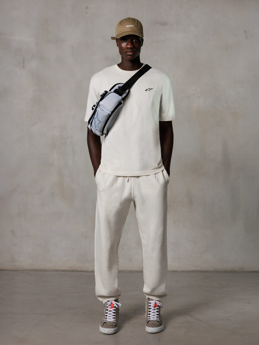 Alpinestars Essentials Ageless SS Tee, Kurzarm-T-Shirt, Off-White, mit einem kleinen schwarzen Logo auf der Brust, getragen mit passender Jogginghose, hellbrauner Baseballkappe und einer grauen Crossbody-Tasche, lässige Lifestyle-Streetwear