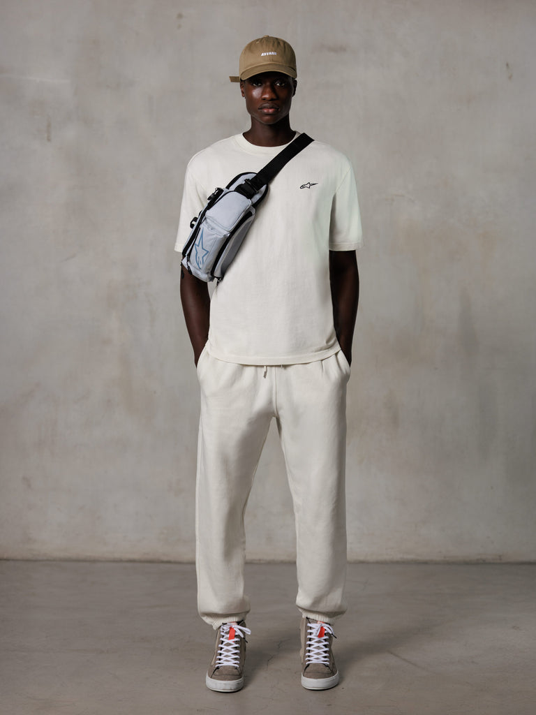 Alpinestars Essentials Ageless SS Tee, Kurzarm-T-Shirt, Off-White, mit einem kleinen schwarzen Logo auf der Brust, getragen mit passender Jogginghose, hellbrauner Baseballkappe und einer grauen Crossbody-Tasche, lässige Lifestyle-Streetwear