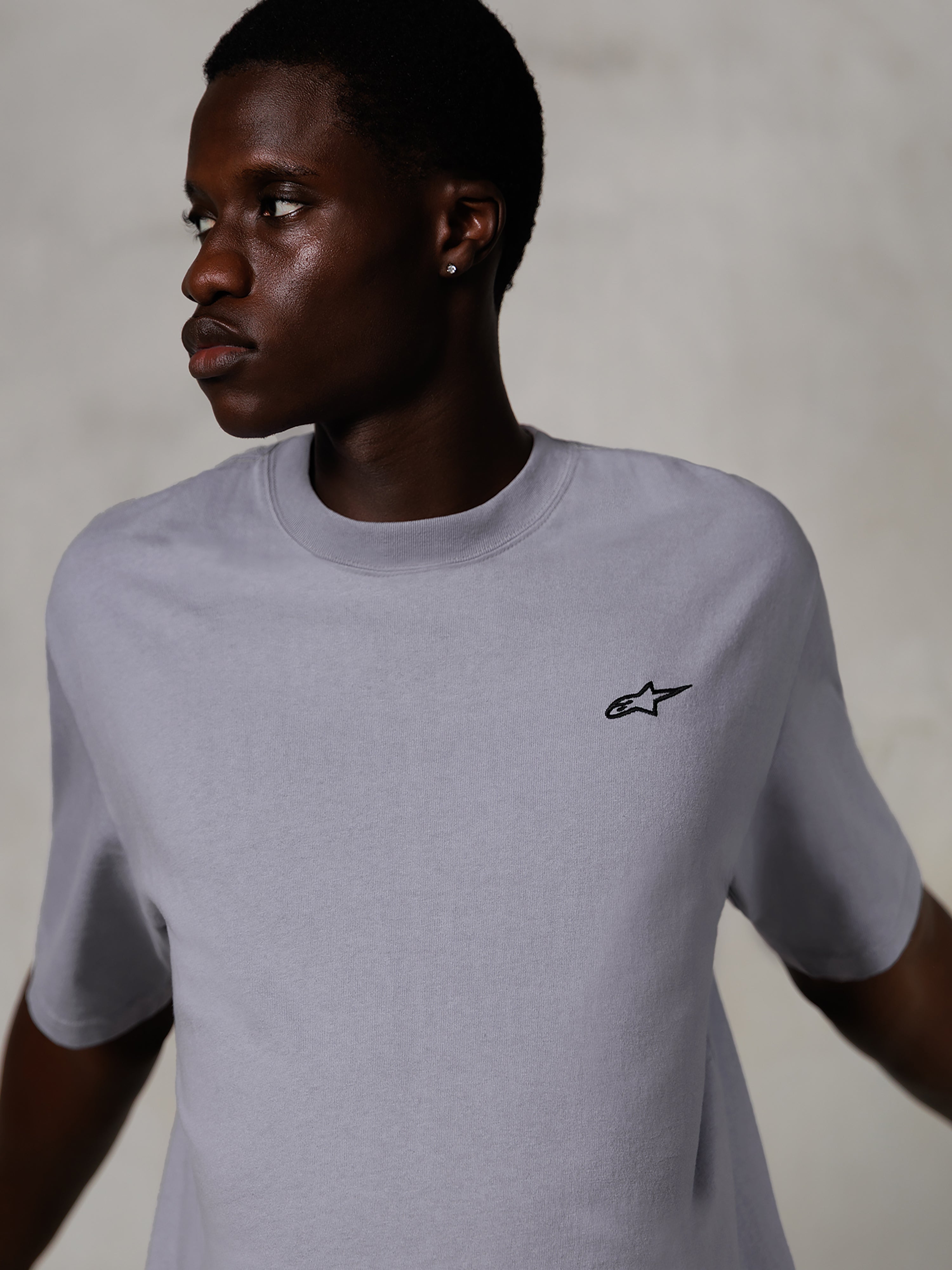 Model trägt das Alpinestars Essentials Ageless SS Tee, kurzärmeliges T-Shirt, Silver, mit einem kleinen schwarzen Logo auf der Brust, entspannte Passform, cleanes und minimales Streetwear-Design