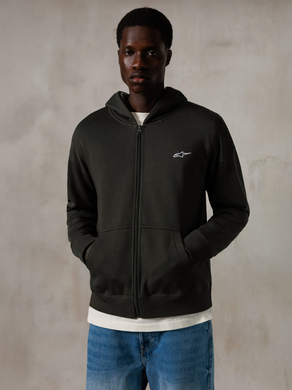 Alpinestars Essentials Ageless Zip Hoodie, Off Black, Zip-Kapuzenjacke mit einem kleinen weißen Alpinestars-Logo auf der Brust, entspannte Passform, getragen von einem Modell mit blauen Jeans und einem weißen T-Shirt, lässige vom Motorsport inspirierte Streetwear