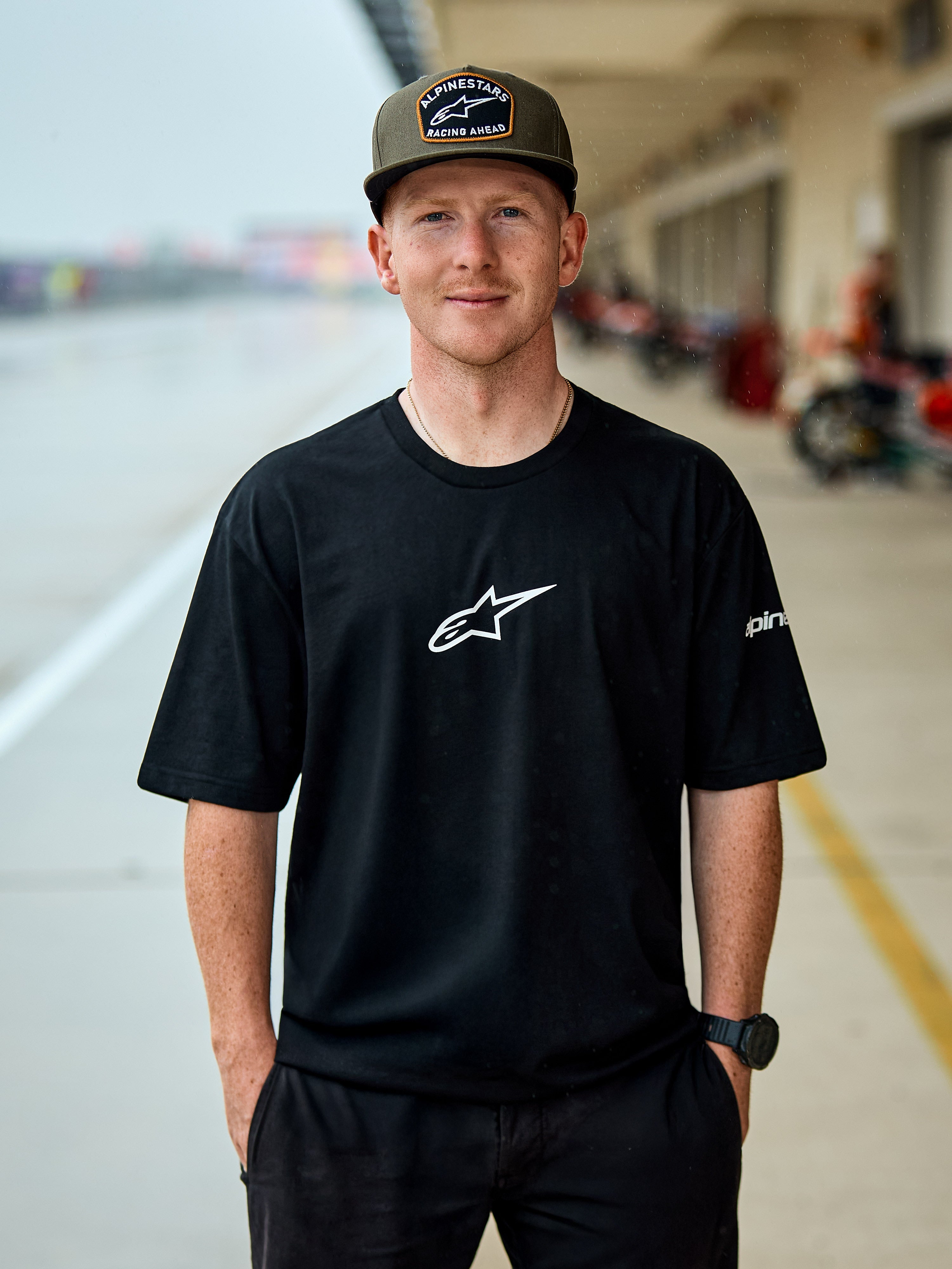 Mann im Alpinestars Frontal CSF Tee, kurzärmeliges T-Shirt, Schwarz, weißes zentrales Astars-Logo, olivgrüne Alpinestars Kappe, lässige vom Motorsport inspirierte Streetwear