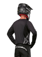 Motocross-Fahrer in der Rückansicht, trägt das Alpinestars 2024 Fluid Graphite Jersey in Black Silver mit einem großen silbernen Logo an der Seite, kombiniert mit einem schwarzen Helm in Carbon-Optik und einer schwarzen Hose, professionelle Offroad-Rennausrüstung