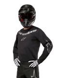 Alpinestars 2024 Fluid Graphite Jersey, Motocross-Trikot in Schwarz und Silber, langärmelige Performance-Passform, getragen mit schwarzem Helm und weißen Handschuhen, silbernes Alpinestars-Branding, Offroad-Rennausrüstung