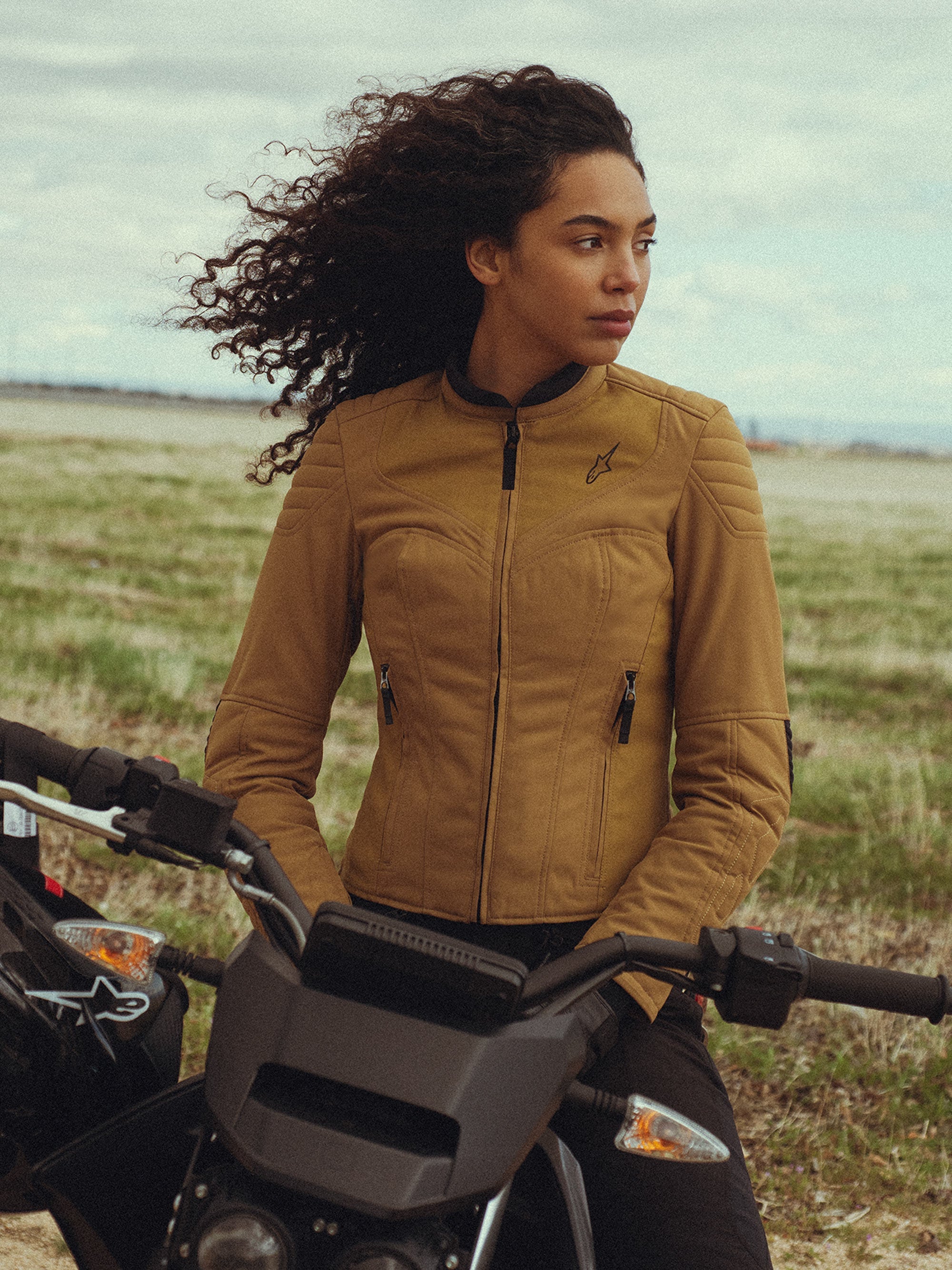 Alpinestars Isla WR Damenjacke in Utility Brown Light Gold, getragen von einer Frau auf einem Motorrad im Freien, mit gesteppten Schulterdetails, schmaler Silhouette und zeitgemäßem urbanem Styling.
