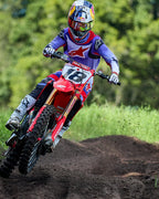 Motocross-Fahrer auf einer roten Honda, Alpinestars 2025 Fluid Wurx Trikot in Lila und leuchtendem Rot, passende Hose, weiße Stiefel und Red Bull Helm, beim Durchfahren einer Kurve auf einer Motocross-Strecke.