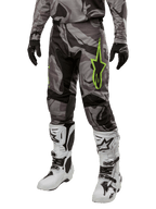 Alpinestars 2024 Racer Tactical Hose, Motocross-Hose, Grau und Schwarz Tarnmuster, neongrünes Stern-Logo, getragen mit weiß-schwarzen Stiefeln und schwarzen Handschuhen, Geländefahrt-Ausrüstung