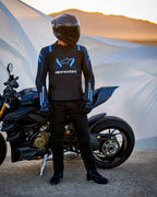 Alpinestars T-Stunt Air Jacke, sportlich geschnittene Textiljacke in Schwarz, Dunkelblau und Hellblau, getragen von einem Fahrer neben einem Ducati-Motorrad, inklusive Carbon-Helm und schwarzer Fahrausrüstung, vor einer Wüstenlandschaft bei Sonnenuntergang.