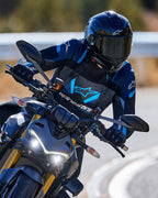 Motorradfahrer in Kurvenlage mit der Alpinestars T-Stunt Air Jacke in Schwarz, Dunkelblau und Hellblau, kombiniert mit schwarzem Helm und Handschuhen, Action-Aufnahme auf einer asphaltierten Straße, Performance-Sport-Fahrerausrüstung