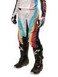 Alpinestars Stella 2024 Techstar Damenhose, Motocross-Hose, Schwarz-Mehrfarbig mit diagonalen roten, gelben und blauen Streifen, getragen mit schwarzen Stiefeln und weißen Handschuhen, mit schützenden schwarzen Knieeinsätzen und markantem Branding für Offroad-Rennen.