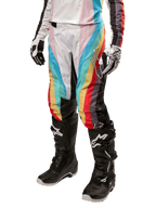 Alpinestars Stella 2024 Techstar Damenhose, Motocross-Hose, Schwarz-Mehrfarbig mit diagonalen roten, gelben und blauen Streifen, getragen mit schwarzen Stiefeln und weißen Handschuhen, mit schützenden schwarzen Knieeinsätzen und markantem Branding für Offroad-Rennen.