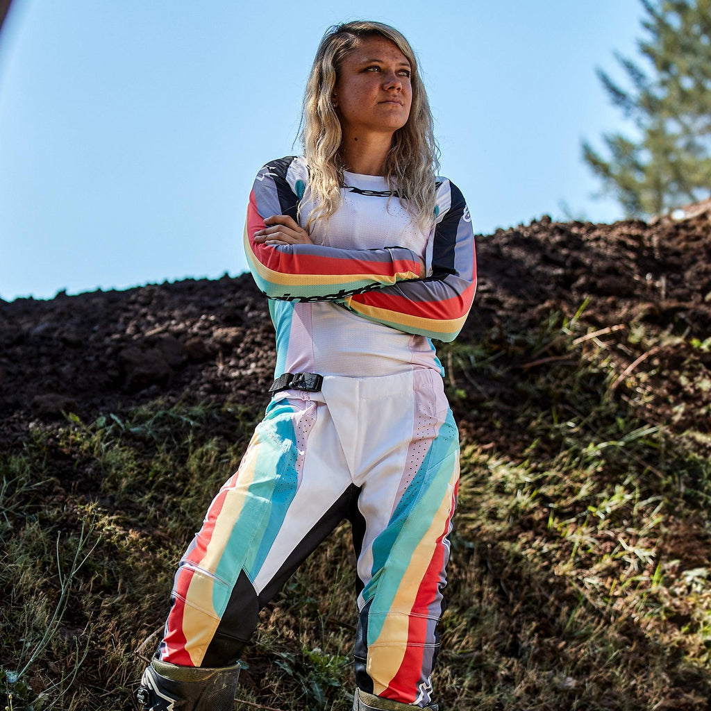 Motocross-Fahrerin in Alpinestars Stella Techstar Damen-Hose 2024 und passendem Trikot, Schwarz Multicolor Design mit weißer Basis und leuchtend blauen, gelben und roten Streifen, stehend mit verschränkten Armen auf einer Motocross-Strecke, professionelles Offroad-Rennoutfit