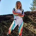 Motocross-Fahrerin in Alpinestars Stella Techstar Damen-Hose 2024 und passendem Trikot, Schwarz Multicolor Design mit weißer Basis und leuchtend blauen, gelben und roten Streifen, stehend mit verschränkten Armen auf einer Motocross-Strecke, professionelles Offroad-Rennoutfit