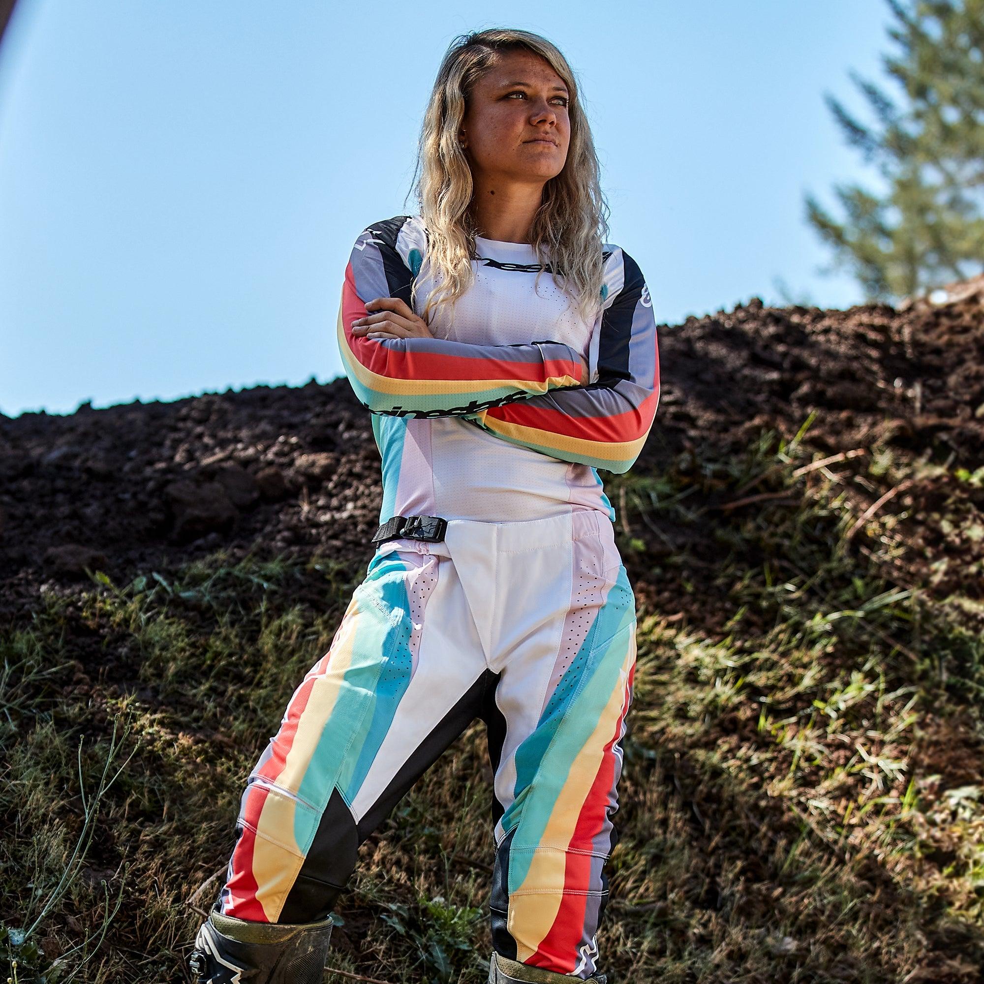Motocross-Fahrerin in Alpinestars Stella Techstar Damen-Hose 2024 und passendem Trikot, Schwarz Multicolor Design mit weißer Basis und leuchtend blauen, gelben und roten Streifen, stehend mit verschränkten Armen auf einer Motocross-Strecke, professionelles Offroad-Rennoutfit
