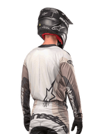 Motocross-Fahrer von hinten im Alpinestars 2024 Techstar Pneuma Jersey in Dark Sand Iron Dust Gray, kombiniert mit einem schwarzen Helm mit Carbon-Finish, mit einem großen Stern-Logo auf dem Rücken und Alpinestars-Branding auf den Ärmeln.