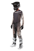 Alpinestars 2024 Techstar Pneuma Hose, Motocross-Hose, Dark Sand Iron Dust Gray, getragen als Teil eines kompletten Outfits mit passendem Trikot, schwarzem Helm, schwarzen Handschuhen und weißen Stiefeln, mit tonalen Wüsten-Camo-Mustern und großen Logo-Akzenten
