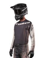 Alpinestars 2024 Techstar Pneuma Jersey, Motocross-Rennsport-Trikot, Dark Sand und Iron Dust Gray, langärmeliges Performance-Oberteil mit lasergeschnittenen Perforationen und weißem Branding, getragen mit passender Hose und schwarzem Helm, athletische Passform für Offroad-Fahrten.