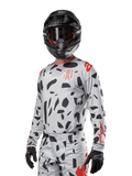 Alpinestars 2024 Techstar Rantera Trikot, Motocross-Trikot, Haze Gray Camo mit schwarzen abstrakten Mustern und roten Akzenten, Langarm-Design getragen mit passendem schwarzem Helm und Handschuhen, technische Offroad-Rennausrüstung