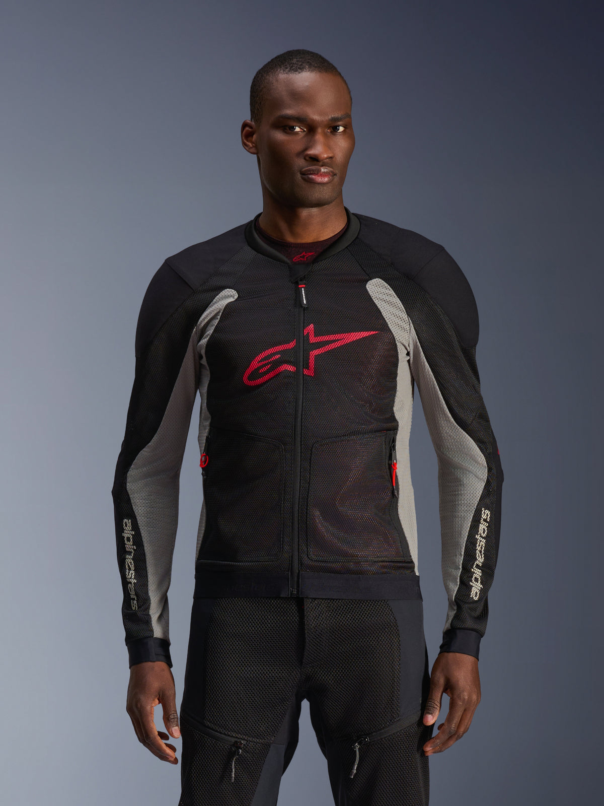 Alpinestars Troop-Air Jacke, Motorrad-Mesh-Jacke, Schwarz und Aschgrau mit leuchtend roten Akzenten, von einem Model getragen, mit atmungsaktiven 2D-Tech-Mesh-Einsätzen zur Belüftung, einem großen roten Astars-Logo auf der Brust und einem technischen, sportlichen Design für den urbanen Straßenverkehr