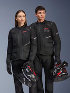 Mann und Frau in Alpinestars Andes Air Drystar® Jacken, schwarze Adventure-Tourenbekleidung mit Mesh-Einsätzen und reflektierenden Details, passende schwarze Hosen und Handschuhe, halten Motocross-Helme, Studioaufnahme von professioneller Motorradbekleidung