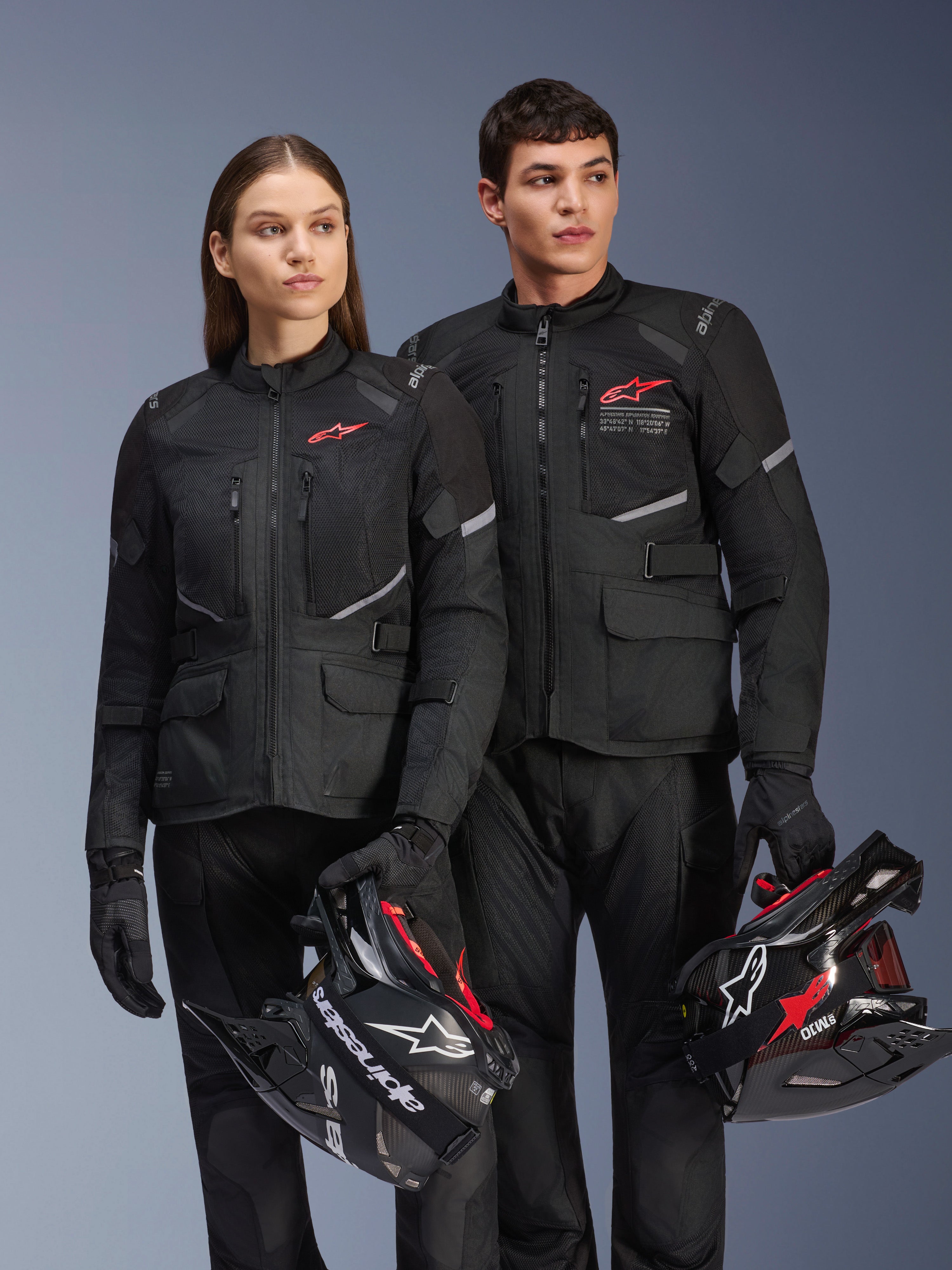 Mann und Frau in Alpinestars Andes Air Drystar® Jacken, schwarze Adventure-Tourenbekleidung mit Mesh-Einsätzen und reflektierenden Details, passende schwarze Hosen und Handschuhe, halten Motocross-Helme, Studioaufnahme von professioneller Motorradbekleidung