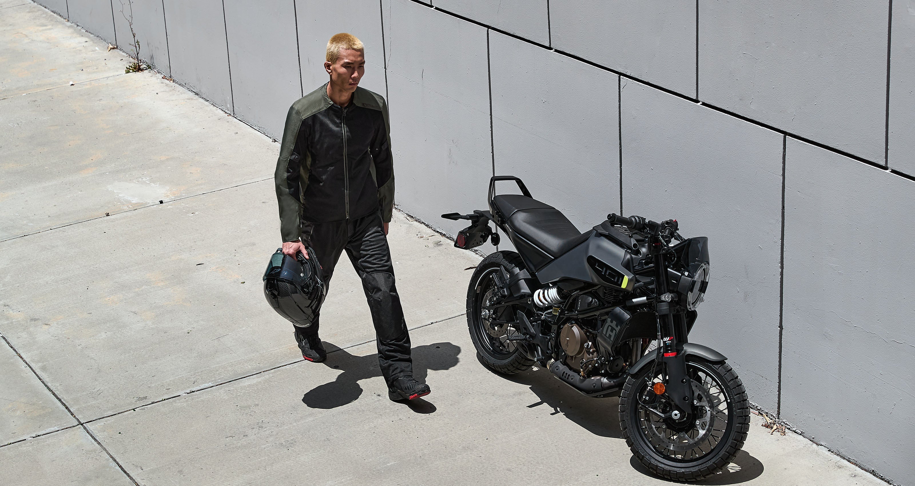 Alpinestars C-1 Air Jacket, urban motorcycle jacket in black and khaki textile, man walking by a bike holding a helmet.